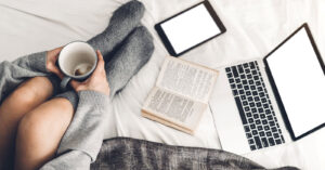 A young adult in grey socks sitting on a bed with a mug surrounded by books & a laptop. Representing someone who could get support in counseling or therapy for college students in Manhattan or Brooklyn. Whether in person or online you can get support finding your path in college in New York today!