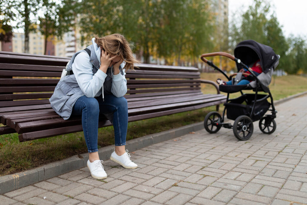 Upset mom with head in her hands sitting next to her baby stroller. Representing someone who needs to talk to a Manhattan postpartum depression therapist. Where she can get support for her maternal mental health by addressing her postpartum anxiety & postpartum depression symptoms.