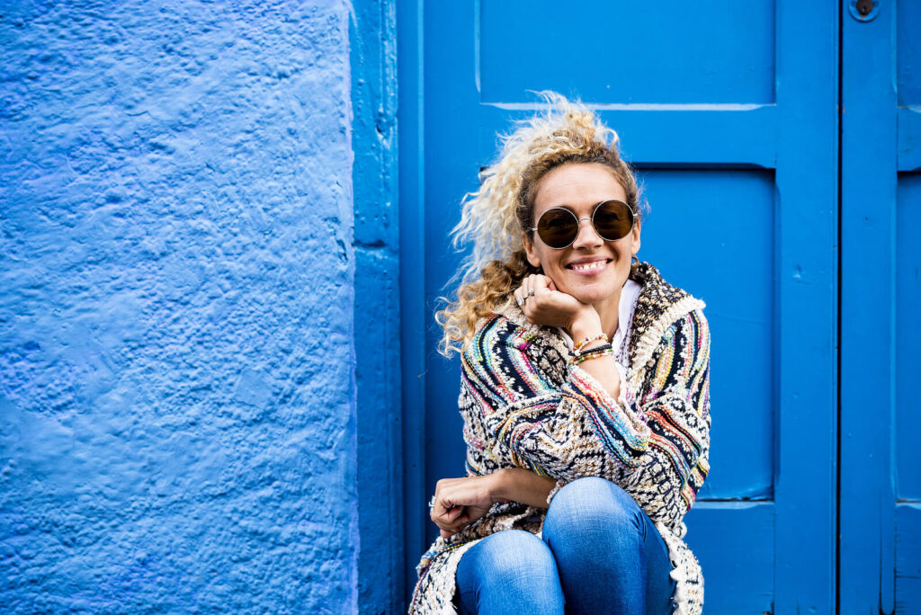 Smiling woman sitting in front of a blue door and wall. Showing the outcome for therapy for women in Brooklyn who are going through a quarter life crisis. With support of a female therapist in manhattan, New York you can feel happy like this.