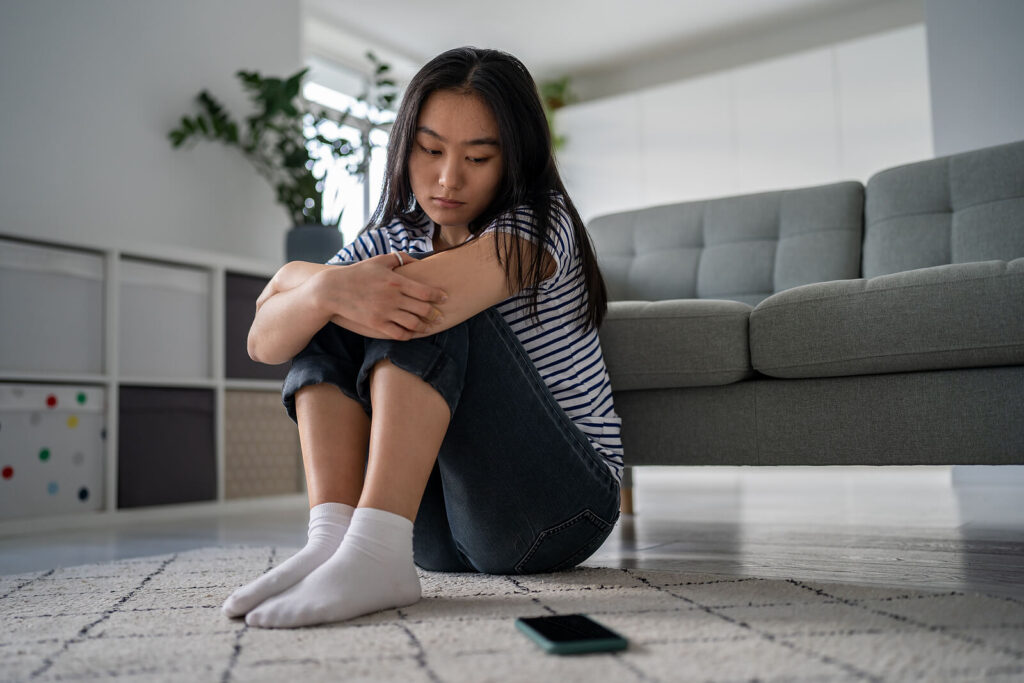 An Asian woman sitting on the ground looking down upset at her phone. Representing someone who could use support from an anxiety therapist in Manhattan, New York. She can start feeling better and stop having panic attacks with anxiety therapy or treatment for social anxiety.