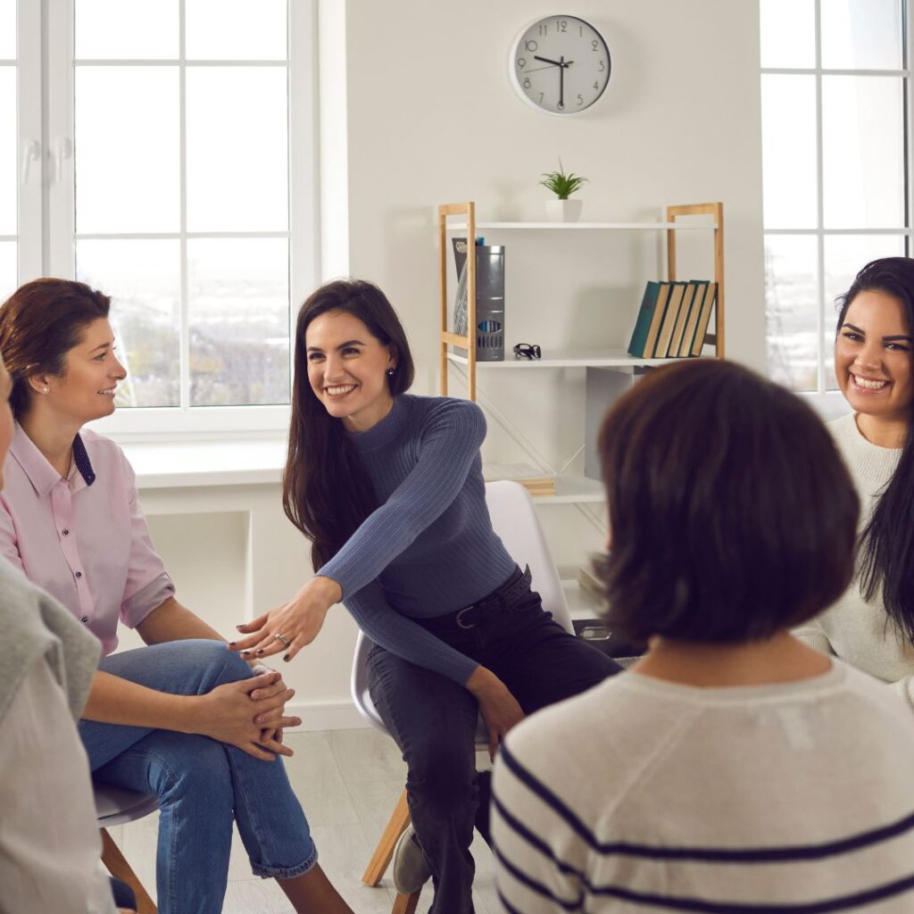 A happy woman sitting in a group of others smiling in an office building in New York. Showing the benefits of anxiety therapist approved tips. You can get anxiety therapy tailored for women in Manhattan, Brooklyn, or throughout NYC. 