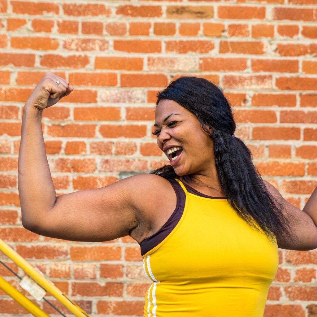 A smiling woman flexes her arm muscles as she workouts out on a  street in New York. Showing the positive benefits of working with a therapist for anxiety treatment for women in Brooklyn & Manhattan, New York.