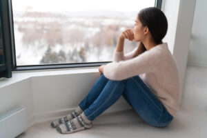 A woman sitting on the floor in front of a window, deep in thought. This represents what depression symptoms can look like during the winter blues. This woman is someone that a Brooklyn & Manhattan depression therapist can help with therapy.