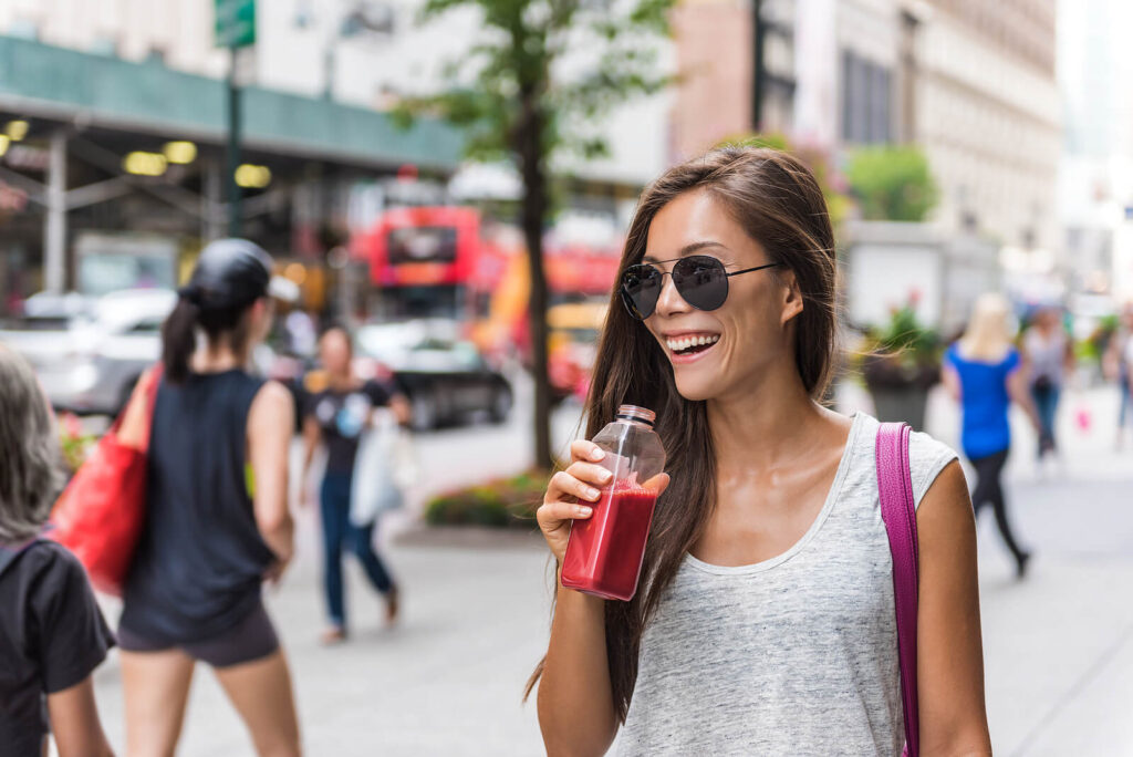 A smiling woman sips on a refreshing juice as she confidently down the street in New York. Showing the positive benefits of CBT for anxiety treatment for women in Brooklyn & Manhattan, New York.