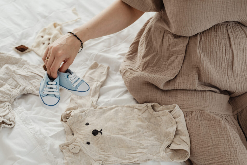 Woman sitting on a bed in Brooklyn holding a pair of baby blue shoes and a bear onsie. Therapy helped her through her fertility journey. 