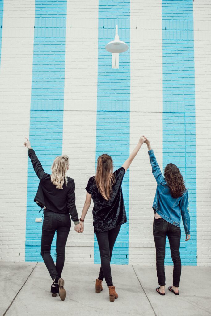 Three friends holding hands in the air in front of a blue stripped wall in Brooklyn. Therapy can help you strengthen your self-esteem and confidence. 