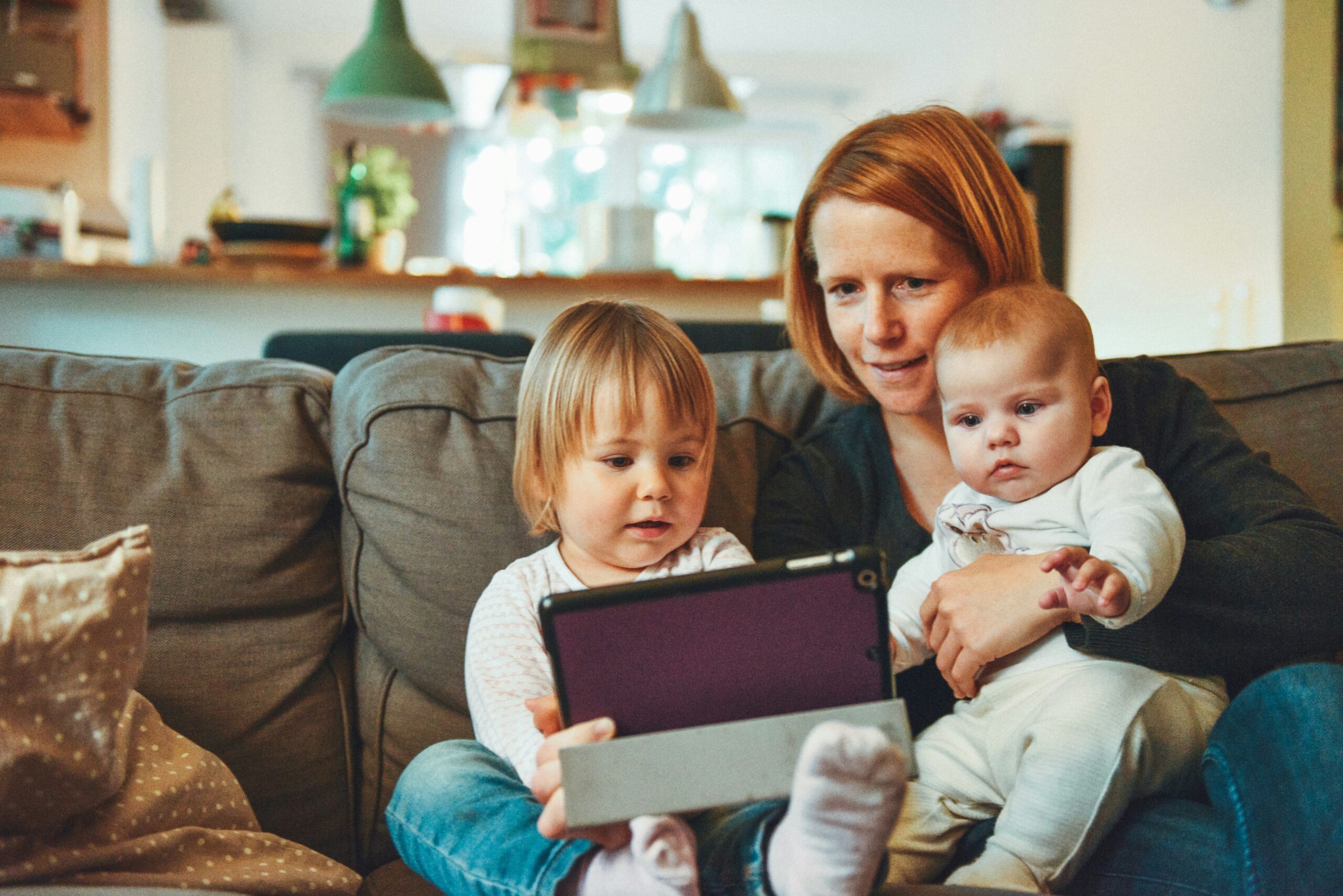 A Brooklyn mother sitting on a couch with her toddler and new baby playing on an ipad. Maternal self esteem is an essential part of maternal mental health.