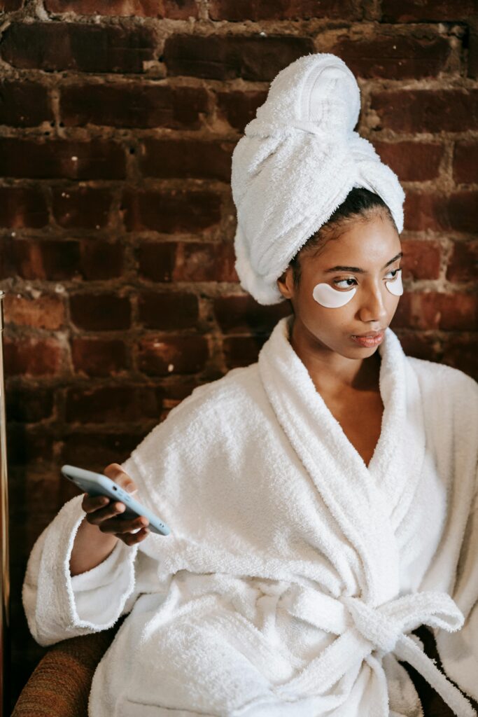 Woman holding her phone with a towel on her hair, robe wrapped around, and eye masks under her eyes. Self care is important for new moms. 