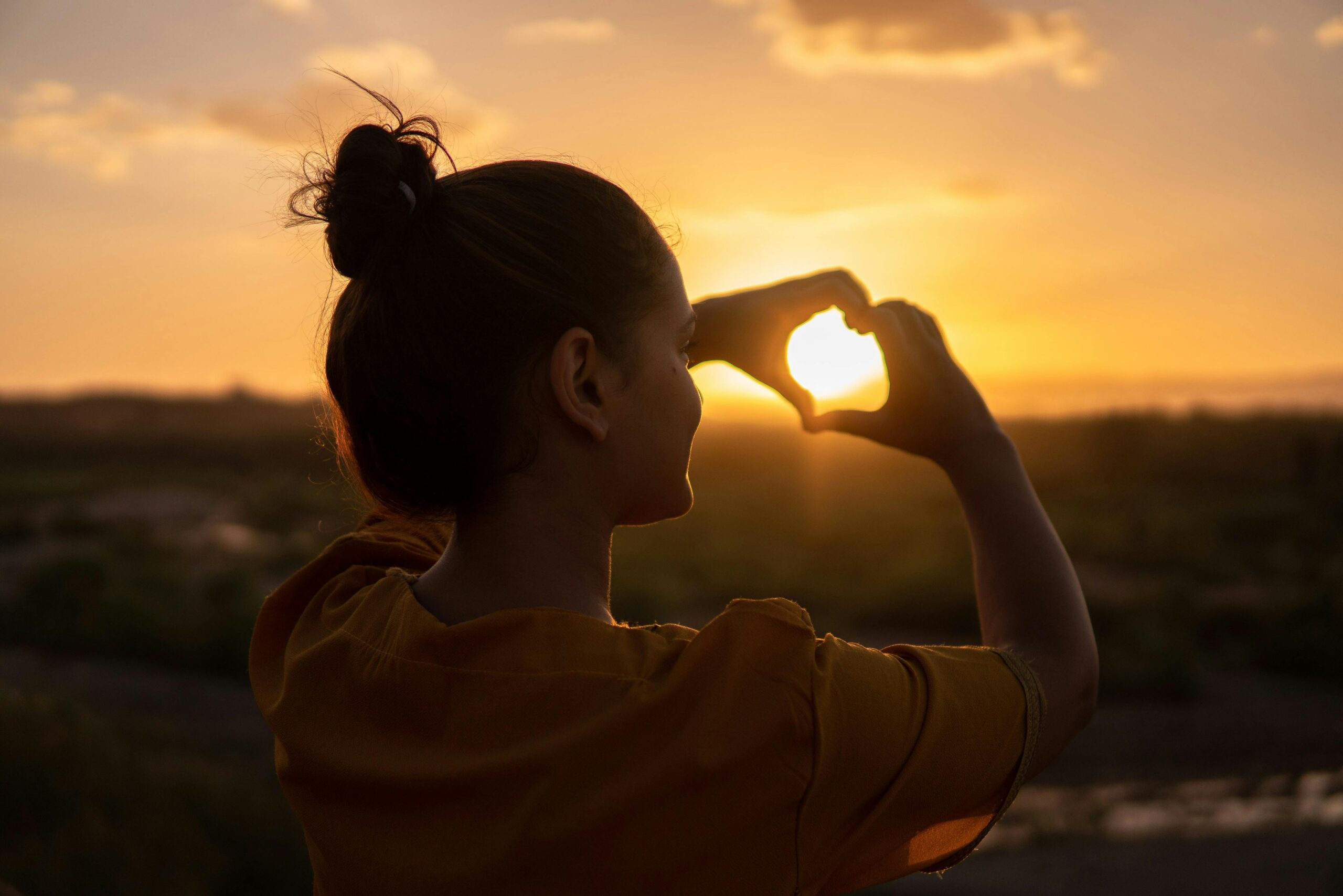 Brooklyn woman, holding her hands in a heart shape with a sunset background. Quieting your critical inner voice is possible. Learn how with our tips and tricks from Manhattan Wellness Therapists.