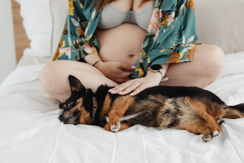 Pregnant mom and dog snuggling on a bed with the woman holding her belly. 