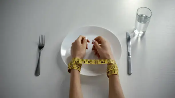 Person's hands wrapped up with a tape measure, over a plate and utensils. Find out more about eating disorders with this blog. 