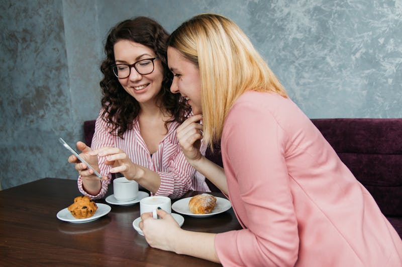 Image of two women getting coffee, looking at a phone both smiling and laughing together.