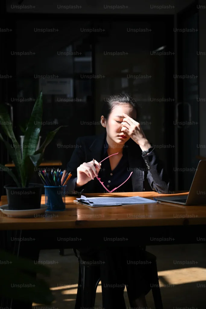 Woman working at desk looking very stressed with one hand on her head and other hand holding her classes. She is working on her stress levels.