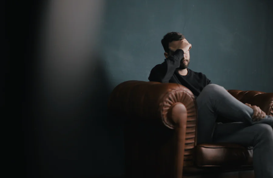 Man sitting on a chair covering his face with his hand and his legs crossed.