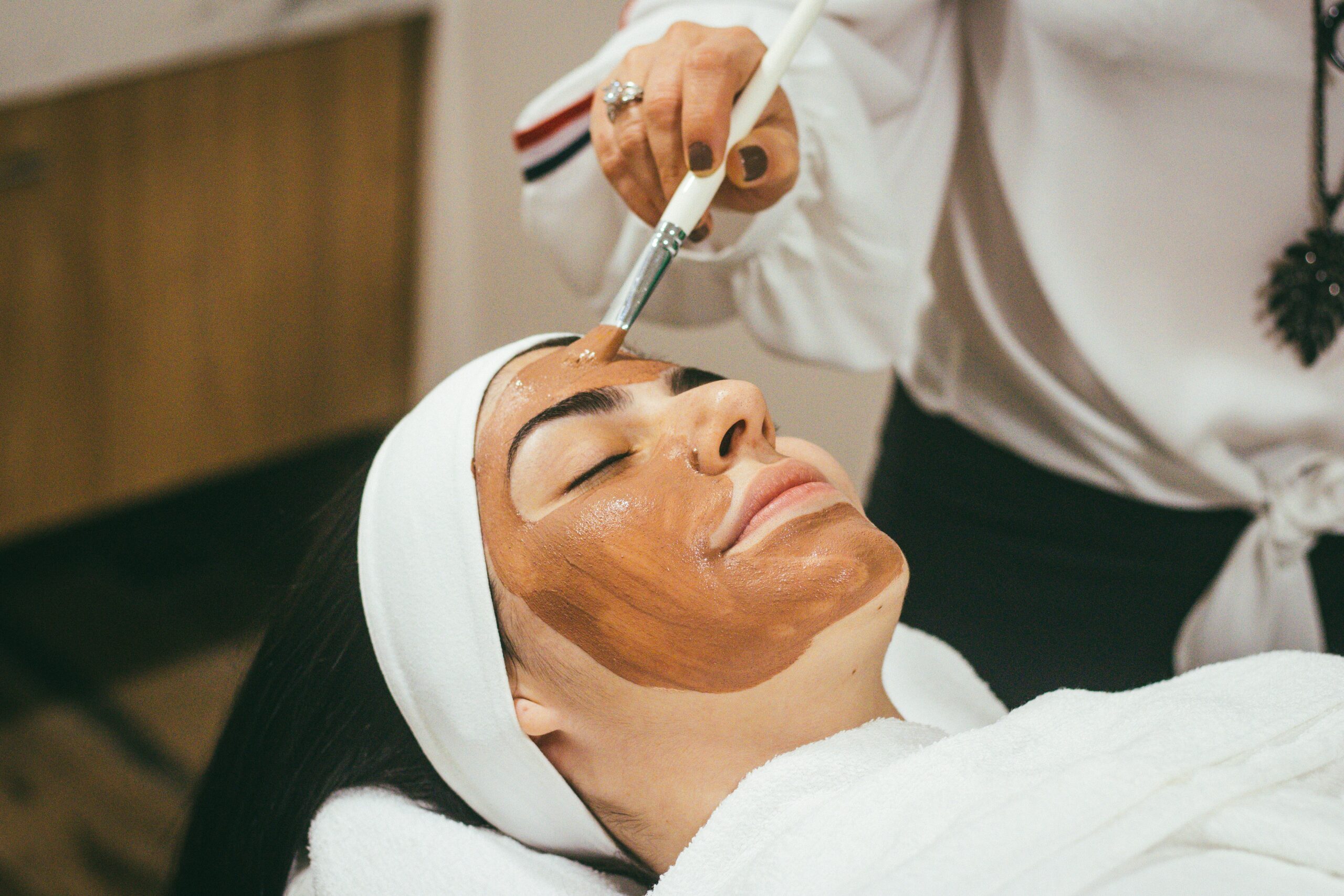 Woman getting a facial with a face mask being brushed on and her eyes closed smiling.