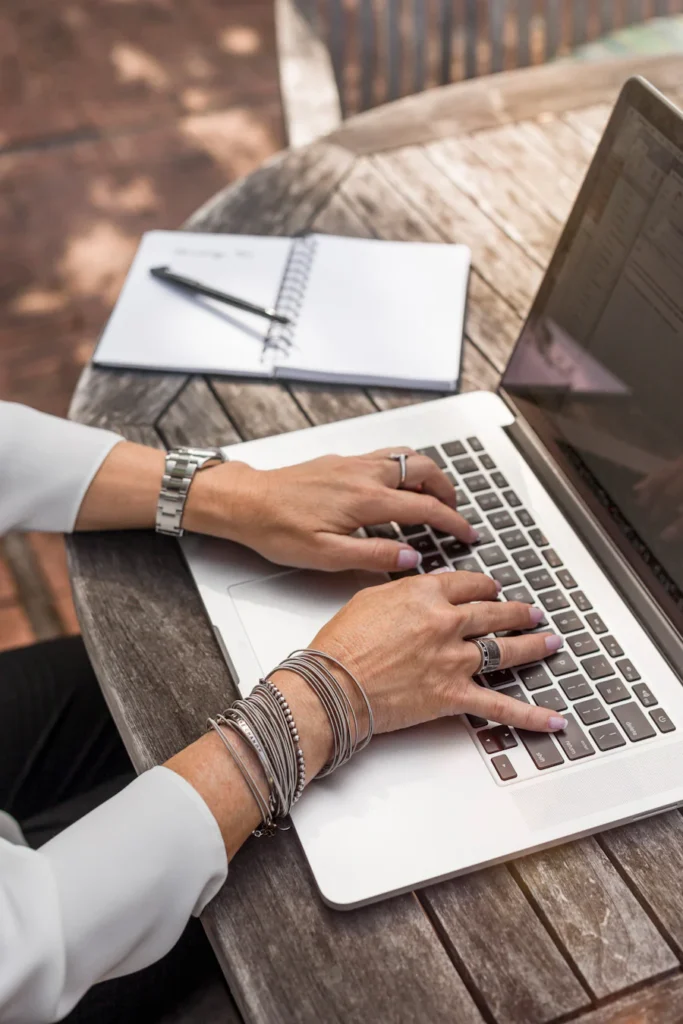 Hands with a lot of silver jewlery typing on laptop on wood table outside.