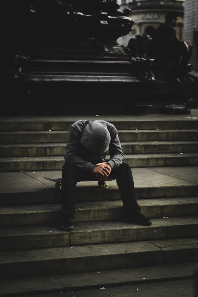 Male sitting on steps on skateboard, looking down with hood on.