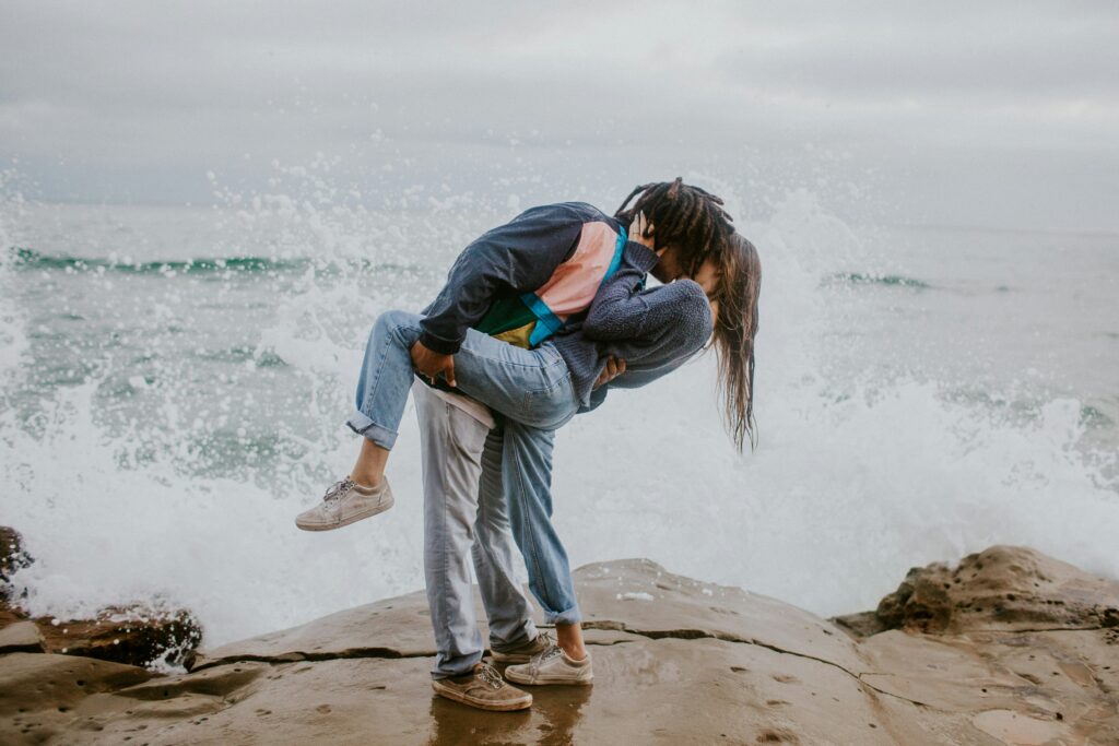 Couple kissing on the beach infront of water.